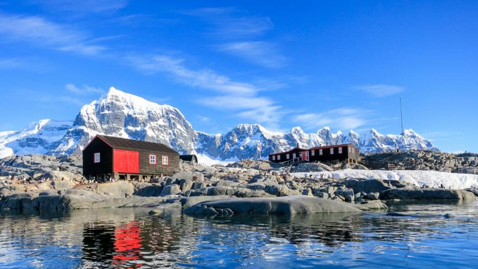 Two red and black huts sit on rocky ground beside calm Antarctic waters, with snow-covered peaks rising in the background. The buildings, part of Port Lockroy, are reflecting in the still surface. Small penguins are also dotted around the landscape and the sky is a bright blue.