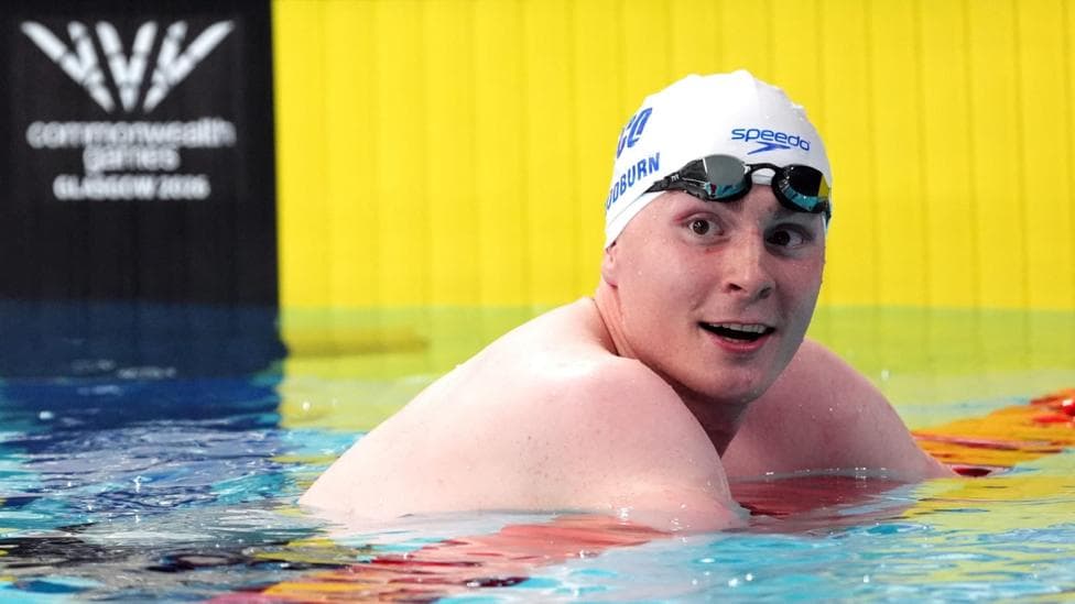 Archie Goodburn following his Men's 50m Breaststroke final at the Tollcross International Swimming Centre during day four of the 2026 Commonwealth Games in Glasgow. He is leaning out of the water on a red lane guide. He is wearing a white swimming cap with blue writing on it and goggles on his forehead.