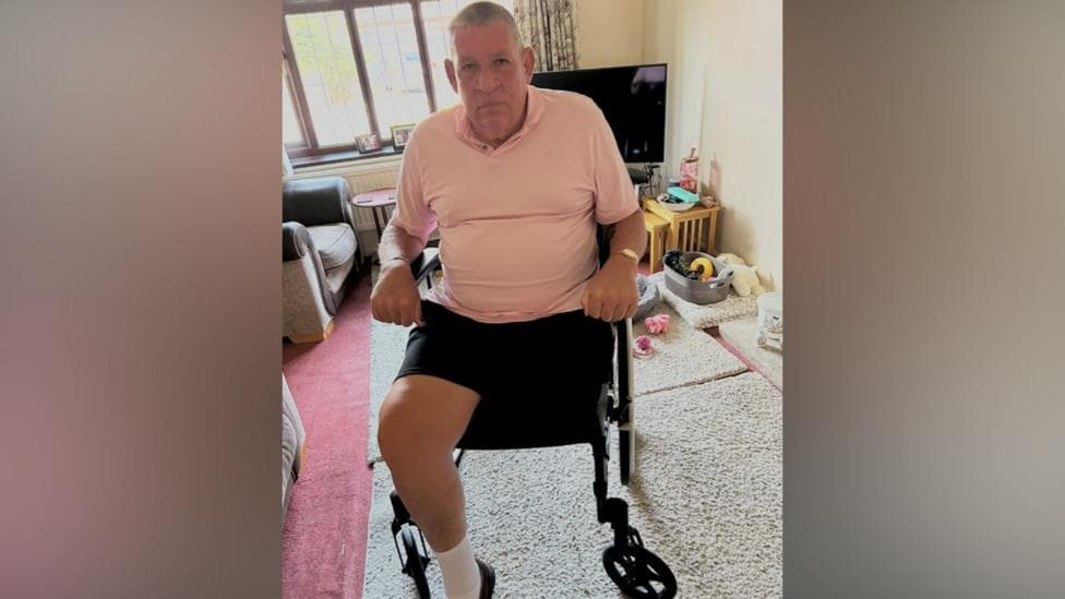 A person sits in a wheelchair in a living room, facing the camera. One leg is visible while the other appears to be amputated above the knee. The room contains armchairs, a television, a rug and a window behind the wheelchair.