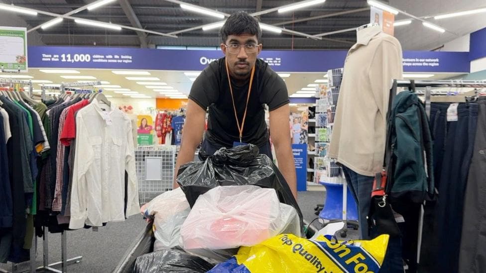 A charity shop worker wears a dark shirt, glasses and an orange lanyard around his neck. He is pushing a trolly of bags through a charity shop