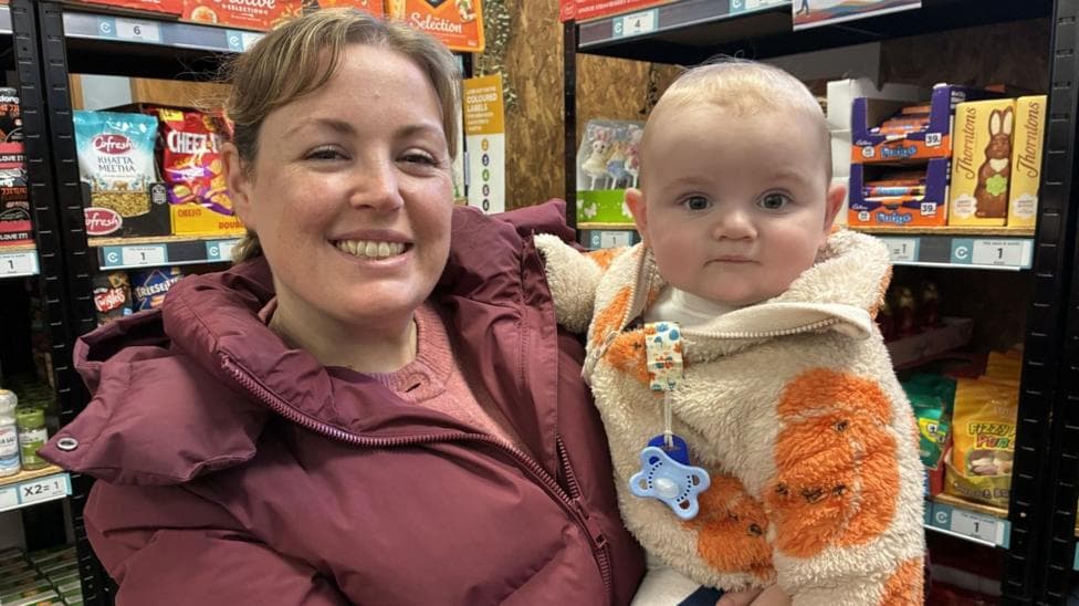 A woman with brown hair tied back in a ponytail is wearing a dark pink padded jacket. She is standing in front of shelves holding a baby with a bald head and wearing a cream fleece with orange pumpkins and a blue dummy