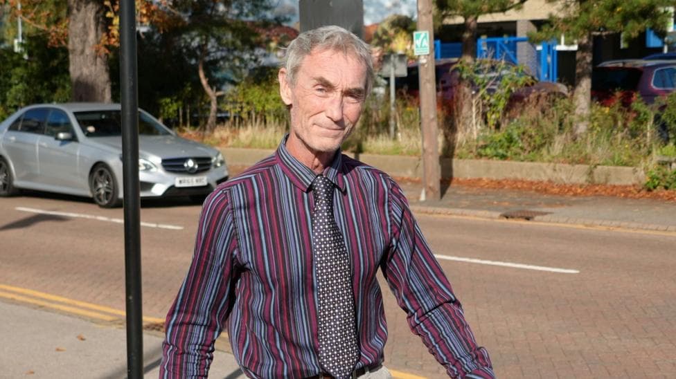 A man smiles at the camera as he walks along a road. He wears a red, blue and purple striped shirt and a polka dot tie. He has short grey hair.