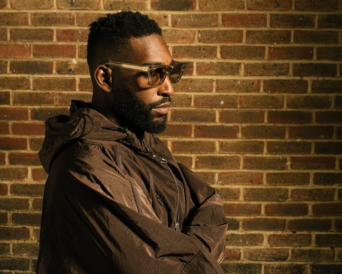 Tinie Tempah wears sunglasses and a brown jacket, arms crossed before a brick wall