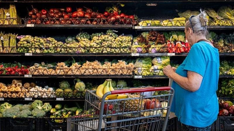 A woman in a blue tshirt stands next to a supermarket trolley as she looks at an aisle stacked with a large variety of vegetables on shelves reaching as high as her head.