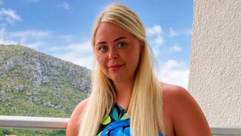A woman with long blonde hair and a flowery dress looking at the camera. She is half-smiling