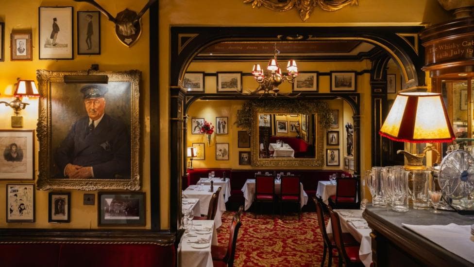 The interior of Rules restaurant. It is a visibly old building with some wooden beams, low light, red and gold carpet, lamps, white table cloths. A painting of an older man in military uniform hangs on the wall to the left of the photo.