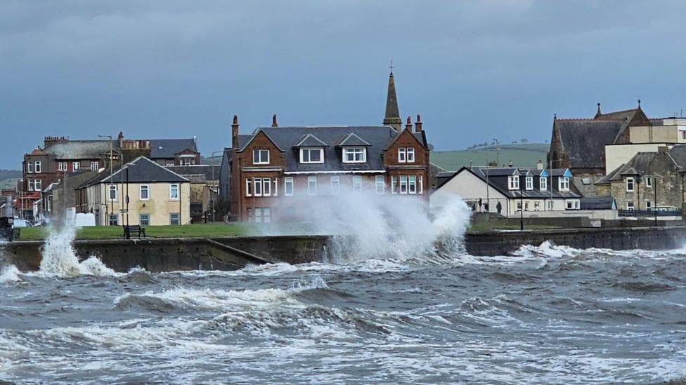 Waves crash against a wall in a coastal town. It is a grey cloudy day and buildings can be seen in the distance