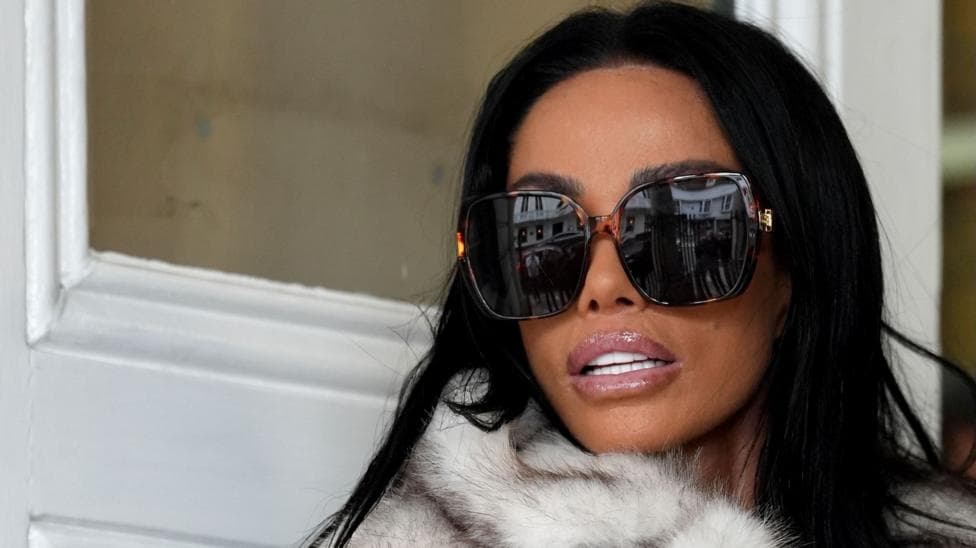 A close up of Katie Price arriving at Lewes Crown Court, she is wearing large dark sunglasses and looking over her shoulder. She has dark hair and is wearing a pale fur jacket, the white wooden doors of the building can be seen behind her.