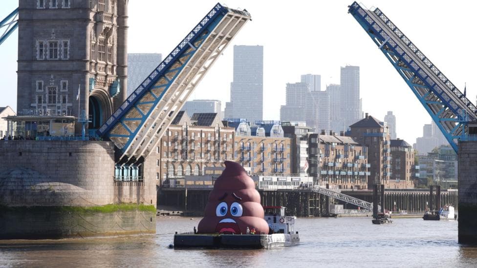 A boat with a large brown poo sculpture sitting atop it. The boat is on the River Thames going under Tower Bridge.