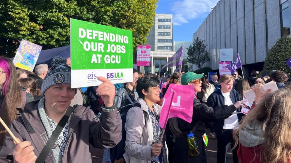 Protestors standing outside a university campus, with many waving placards or flags. One person close to the camera holds up a large green and white sign saying DEFENDING OUR JOBS AT GCU, with others hold pink or purple flags or banners.