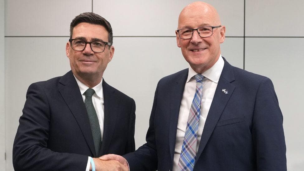 Andy Burnham, who has dark hair and glasses, and John Swinney, who is bald with glasses, shake hands while smiling for the camera