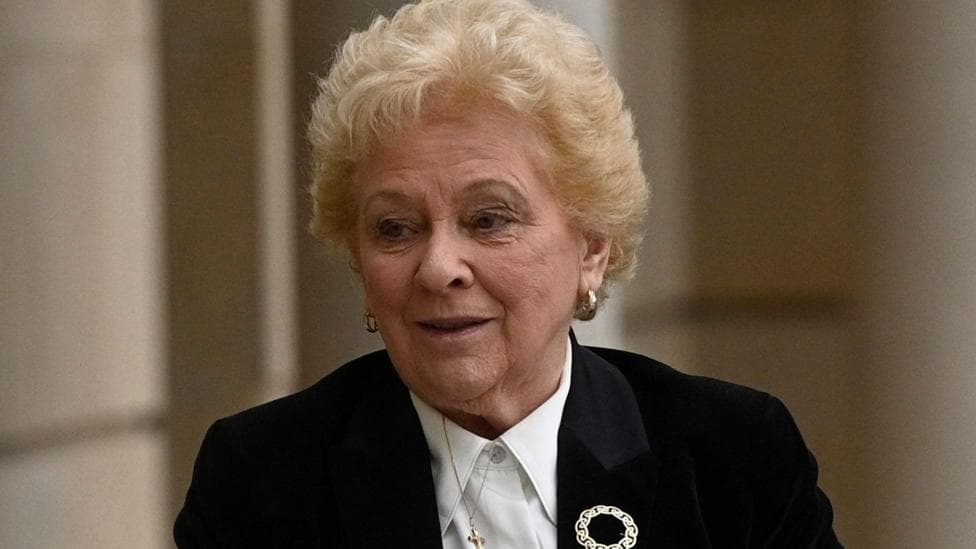 Baroness Nuala O'Loan - a woman with short, curly blonde hair, pictured outside a tall building. She is wearing a black suit jacket over a white blouse, a silver brooch and gold hooped earrings.