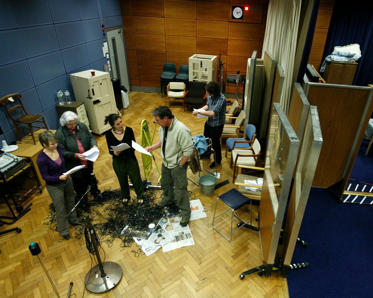 A group of actors holding scripts stand in a huddle in a studio filled with equipment