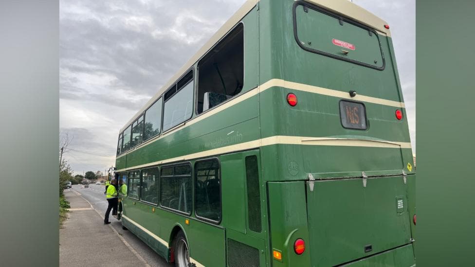 A green double-decker bus with the top right-hand window panel missing. There is a person in high-visibility jacket entering the bus via the front door.