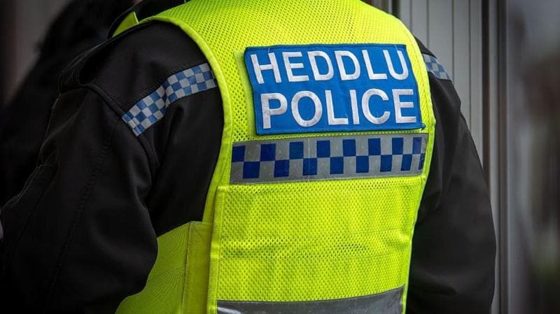 A close-up of a police officer's uniform. They wear a black jacket and yellow high-vis vest which reads: "Heddlu, Police".