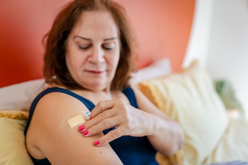 A senior woman wearing a blue vest sits on a bed while applying a hormone replacement therapy patch to her arm.