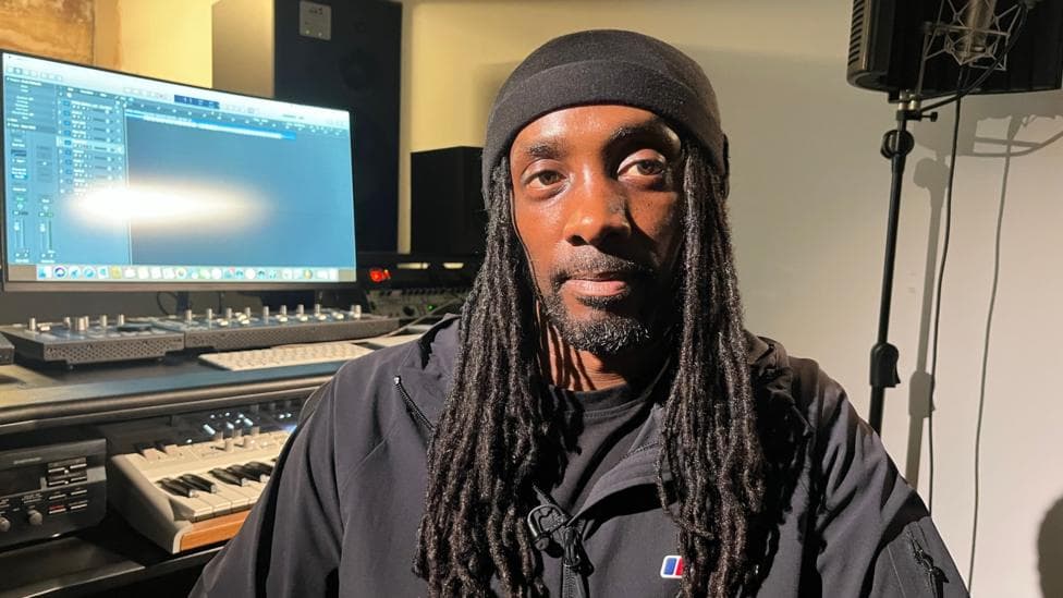 Zimbo is wearing a black hat and has long dreadlocks and a black jacket. Behind him is a computer screen and recording equipment.