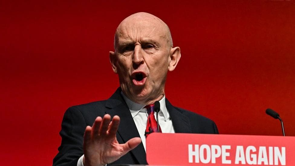 Chancellor John Healey wears a dark suit, white shirt and red tie as he addresses Labour conference attendees behind a podium bearing the party's "hope again" branding.