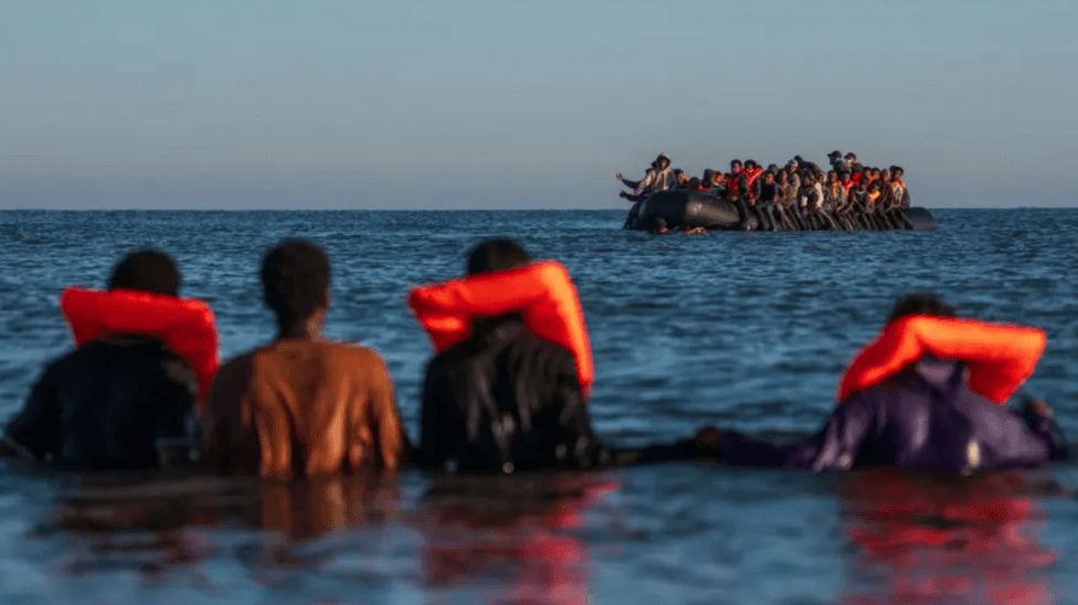 A group of migrants are seen in the distance on an inflatable boat in the Channel. In the foreground are four people in the sea wearing life jackets.