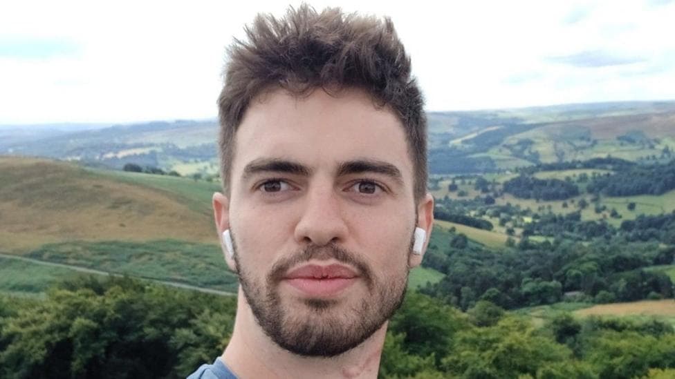 A selfie of William Chapman outdoors, with rolling hills, woodland and patchwork fields stretching across the landscape behind him. He is wearing wireless earbuds and is standing in front of a panoramic countryside view under a bright, cloudy sky.