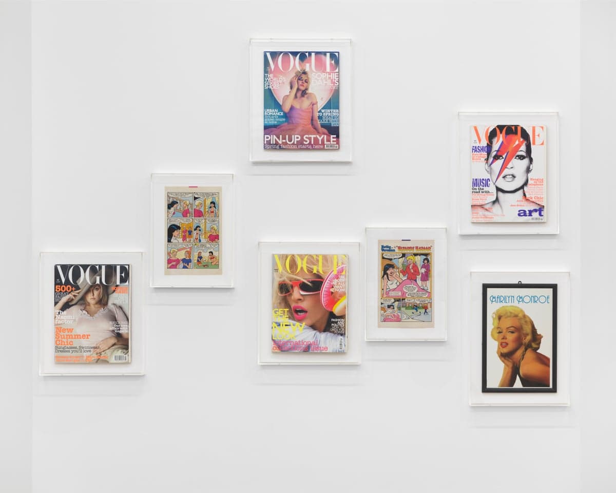 Framed magazine covers and comic pages, including Vogue issues and Marilyn Monroe, hang on a white wall