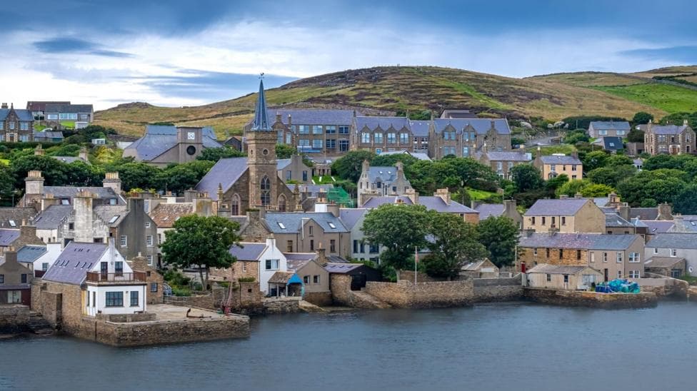 Elevated view across Stromness, Orkney, showing a cluster of closely packed grey and white buildings with pitched roofs, the town hall prominent in the centre, and the surrounding streets and buildings extending across the hillside.