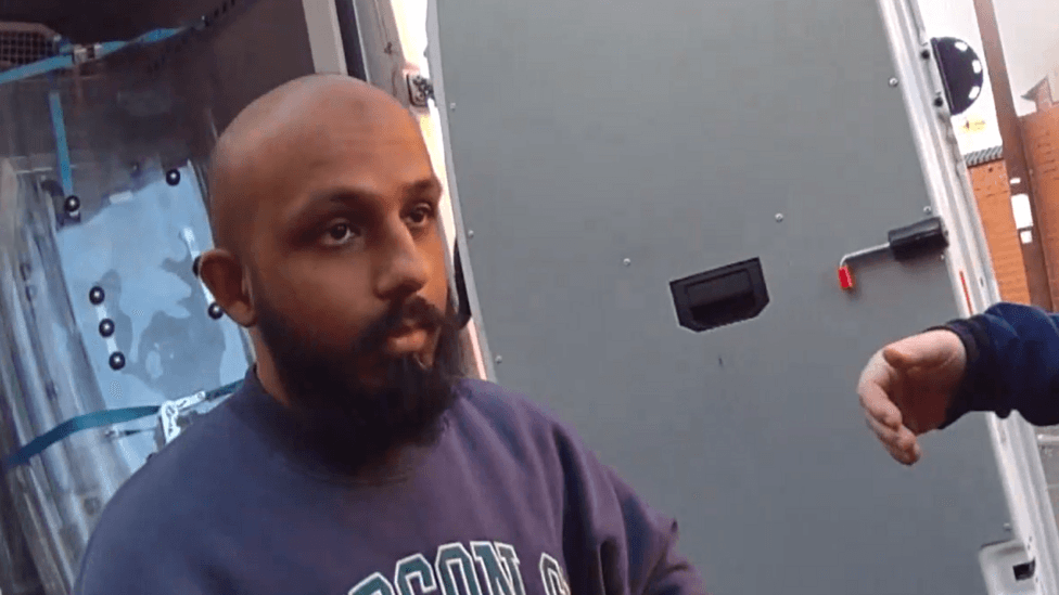 Abusali Rahman, a bald man with a dark beard wearing a grey sweatshirt, stands beside an open vehicle door. The image is taken from police body-worn camera footage.