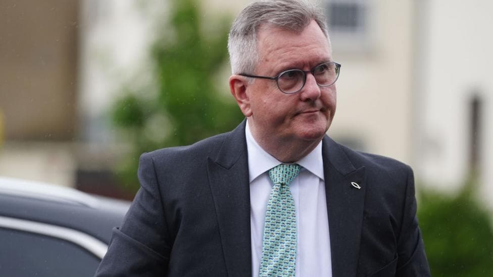 A man with grey hair and glasses wearing a white shirt, green tie and a dark jacket