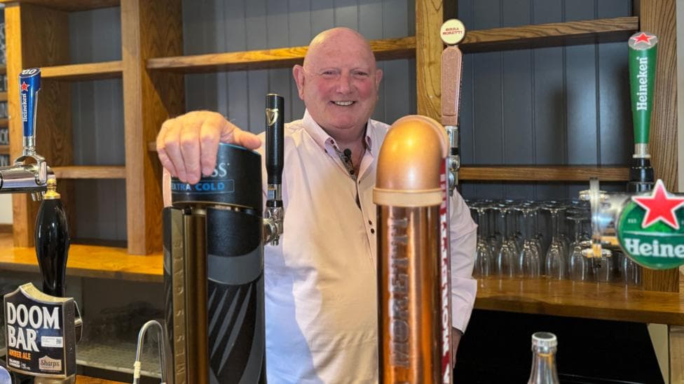 Landlord Patrick Hughes is stood behind the bar in his pub. He is wearing a pale pink shirt.