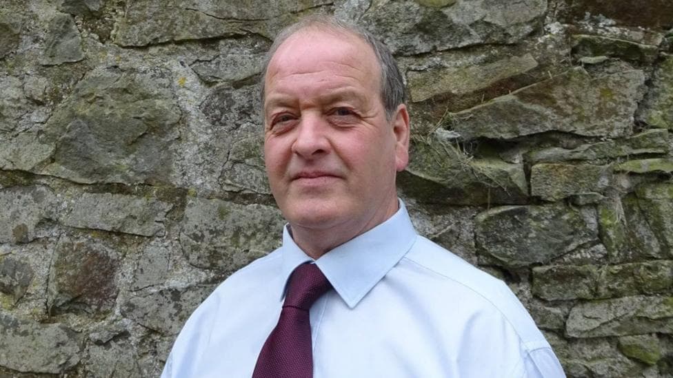 Alan Watson wearing a burgundy tie and a light blue shirt. He stands in front of a stone wall and looks at the camera. He has grey short hair and blue eyes.