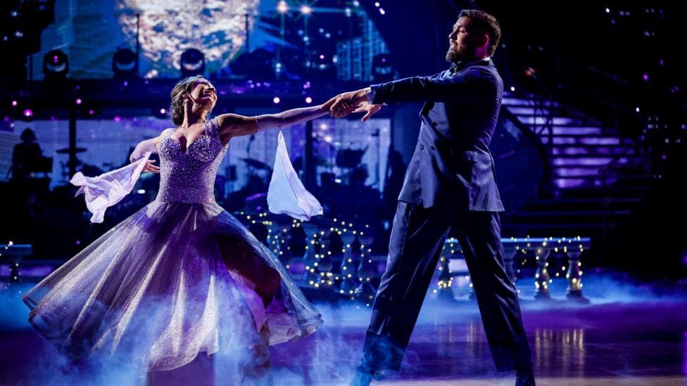A couple on the dance floor during a routine where their arms are outstretched. The woman is wearing a purple ballgown and the man a purple suit. There is a stairwell in the backgroud and lots of fairy lights.