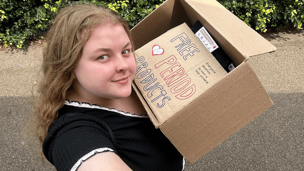 Molly in a selfie photo. She has long wavy light-brown hair, wearing a black top with white trim and holds a cardboard box containing packets and a handwritten sign reading "Free Period Products".