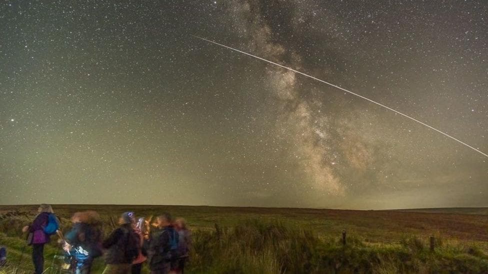 A group, intentionally blurred by the long exposure of the camera, is walking out of frame on the bottom left of the picture. They are walking through a green, hilly landscape in the dark. In the sky, hundreds, if not thousands, of stars shine, while a bright line shoots across frame from the right into the middle of the sky.