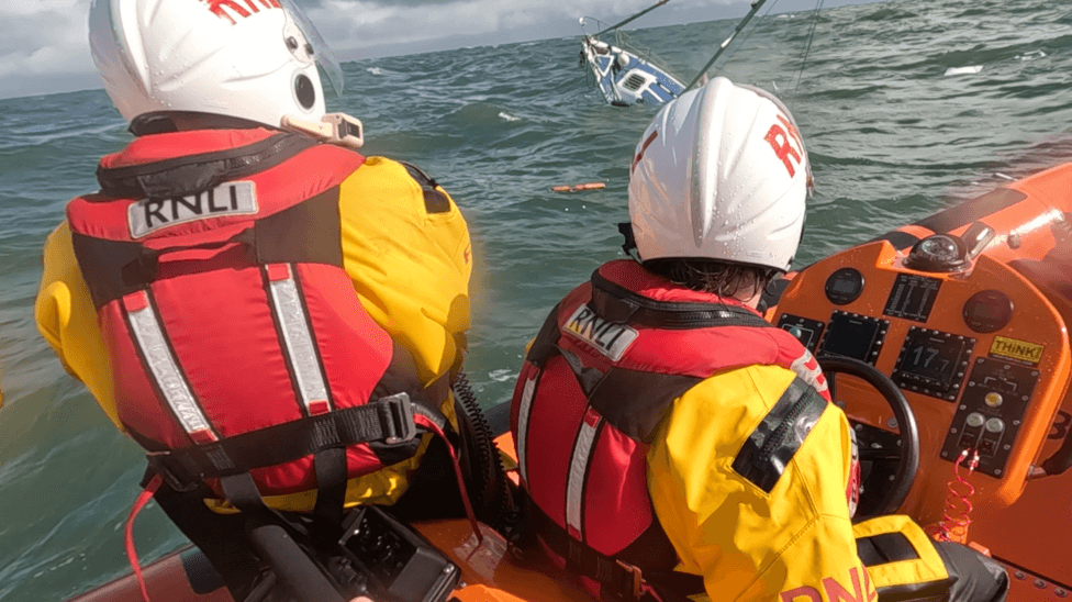 RNLI lifeboat crews on a boat wearing yellow jackets, red lifejackets and white helmets. There is a boat that is sinking in the background.