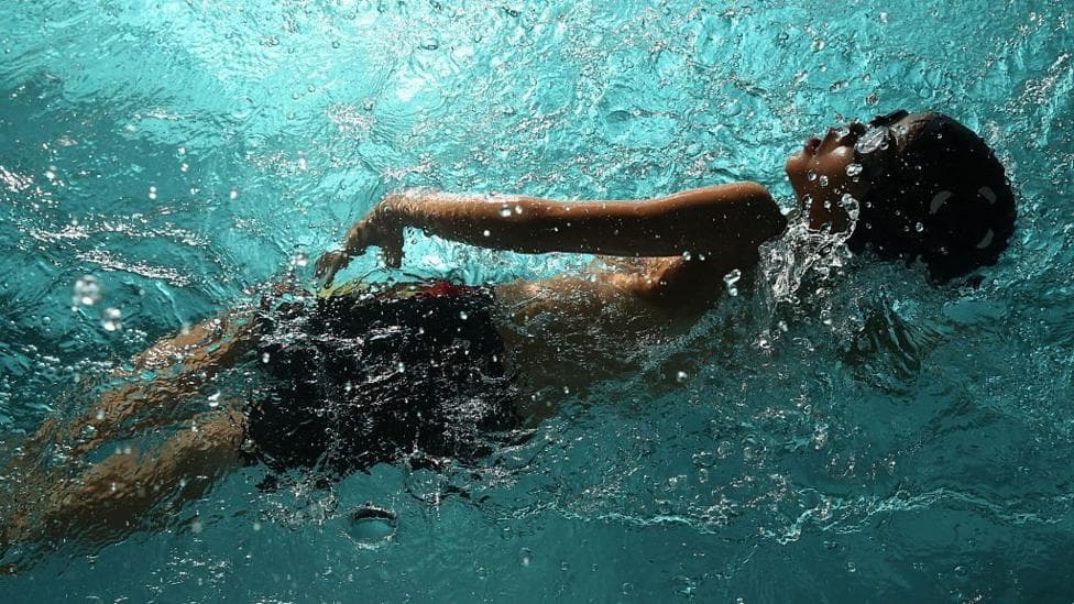 A young boy wearing googles pictured in a pool, taken from below the water. The water goes from crystal blue to a shadowy grey-blue.