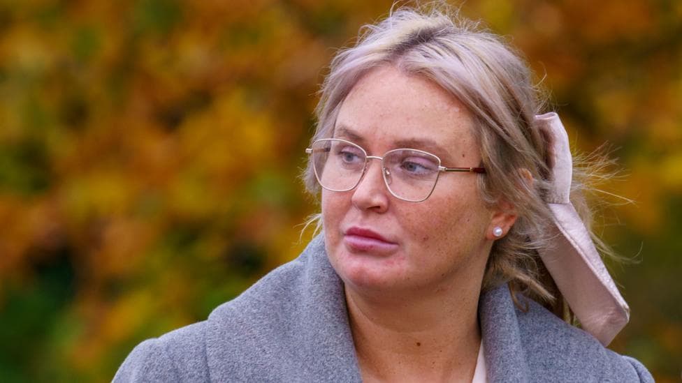 Julie Ann McIlwaine, who has blonde hair tied back in a light pink bow. She wears gold-rimmed glasses and a grey coat.