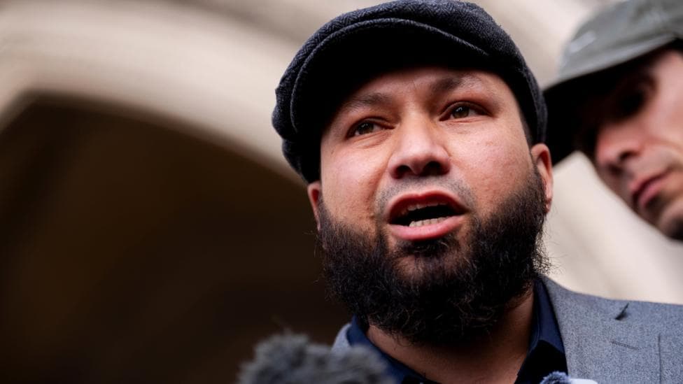 A man wearing a flat cap stands outside a court building and addresses members of the press.