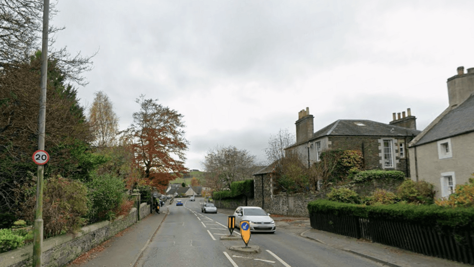 The road through Innerleithen with a 20mph sign on it and cars passing through between houses and trees