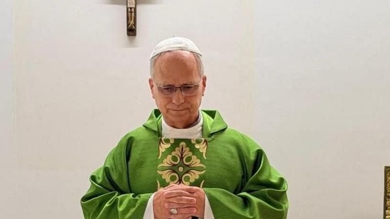Pope Leo celebrates mass from hospital, wearing a green robe and standing before a white wall adorned with a wooden crucifix.