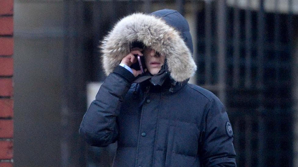 A man wearing a fur hooded black jacket covering his face with a phone held up to his ear.