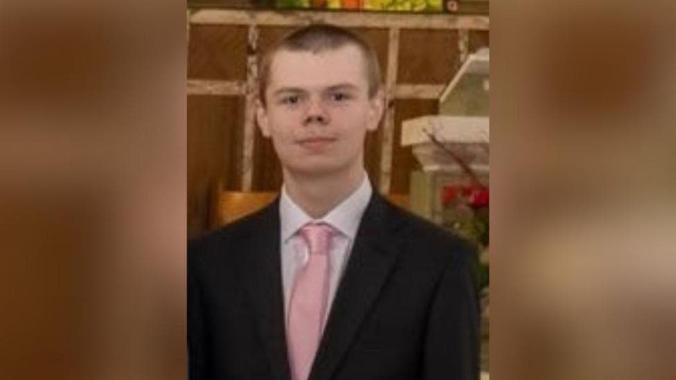 Jack Mellon is seen with with short brown hair, wearing a white dress shirt, a light pink tie, and a black suit jacket, posing indoors against a warm background.