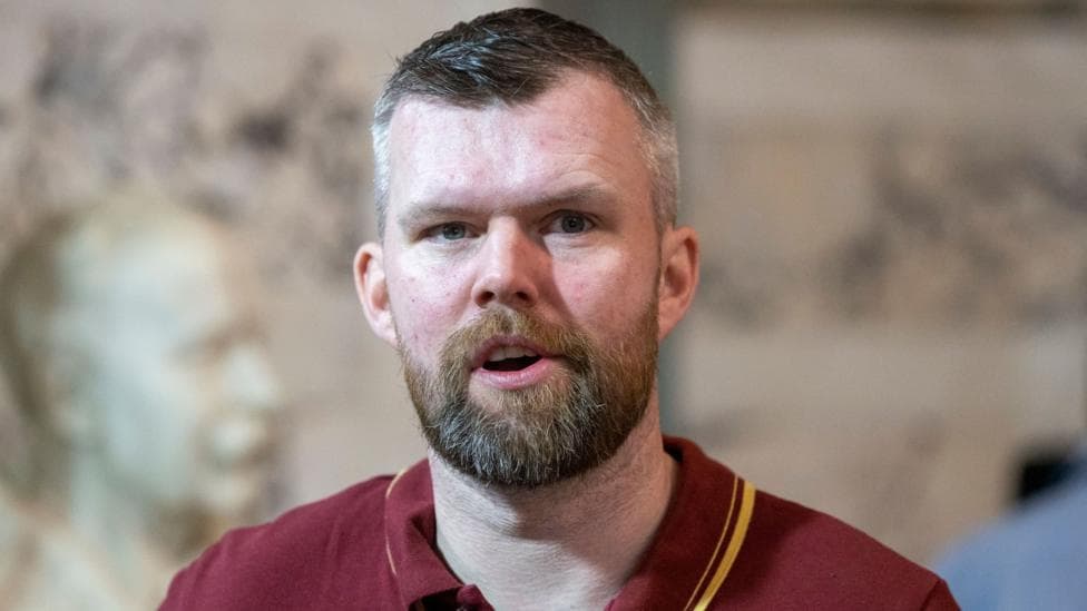 Carroll has short dark hair and a brown beard and moustached. He is wearing a red polo shirt with a yellow line on it.