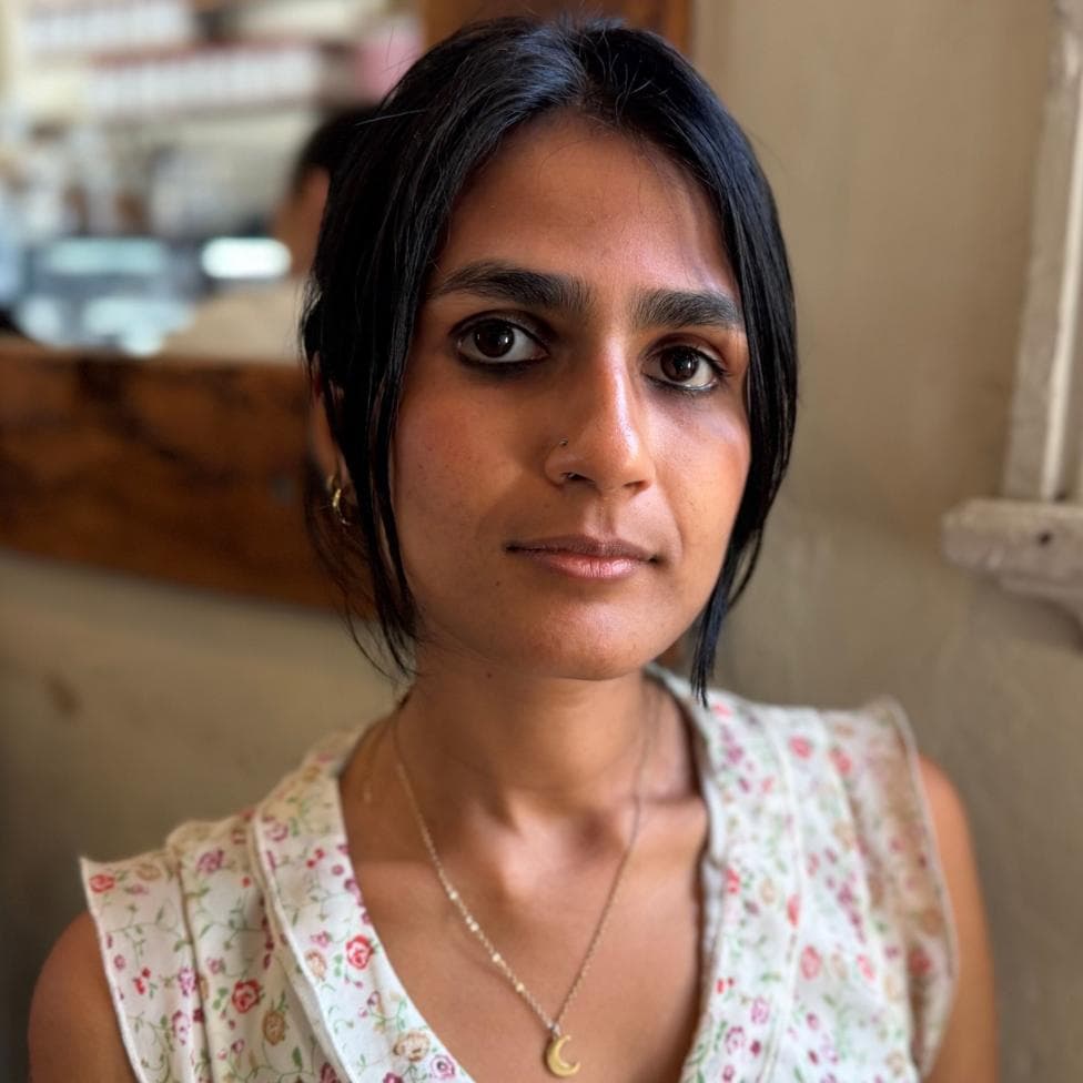 Varsha, wearing a white floral top and moon necklace, looks at the camera