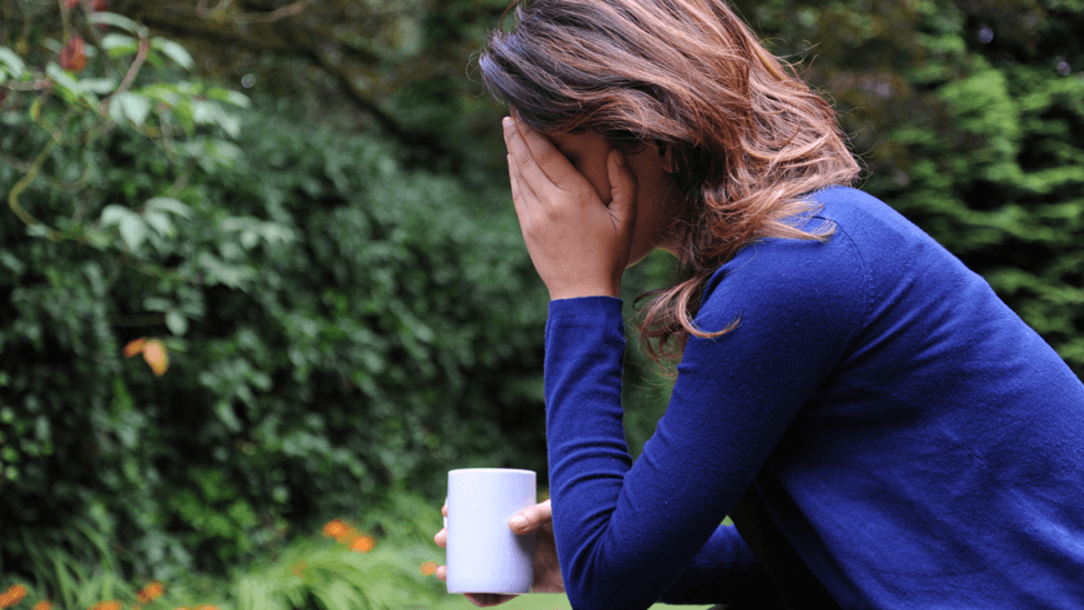 A woman with dark, shoulder-length hair wearing a dark blue jumper sits in a garden with her elbow resting on her knee and her head in her hand. Her face is obscured by her hand and in her other hand she holds a white mug.