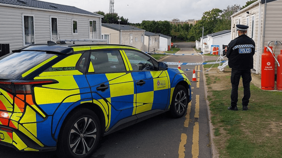 A police car parked on double yellow lines outside a caravan park. A policeman is standing next to the car with their back to the camera. The car is parked in front of police tape and a caravan site