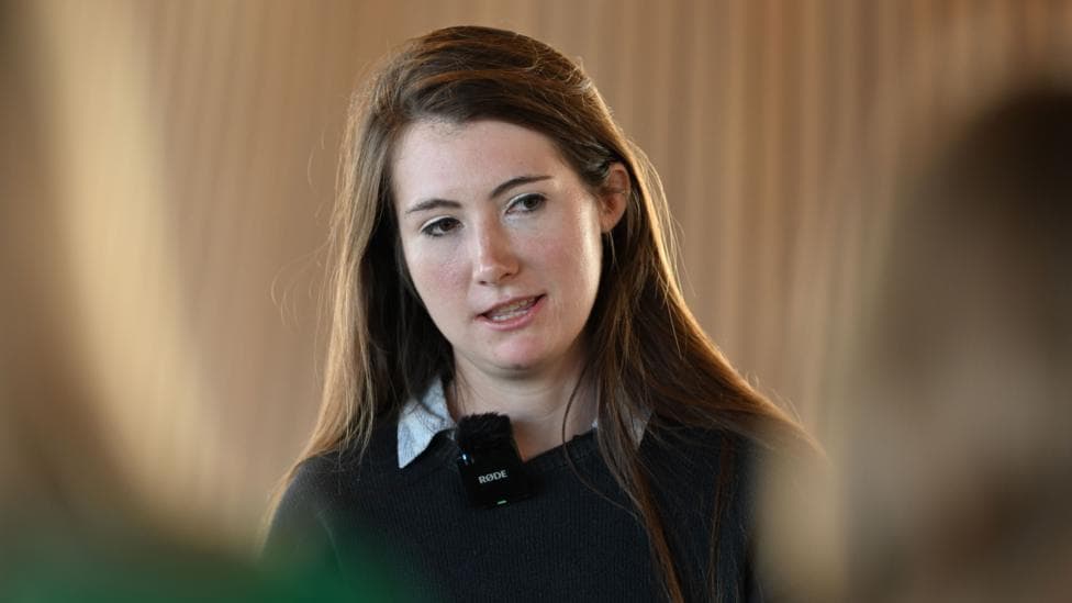 Sarah Cooper-Lesadd. She has dark hair going past hair shoulders in a side parting going. she has dark eyes and eyebrows. she is wearing a black jumper with a white collared shirt underneath