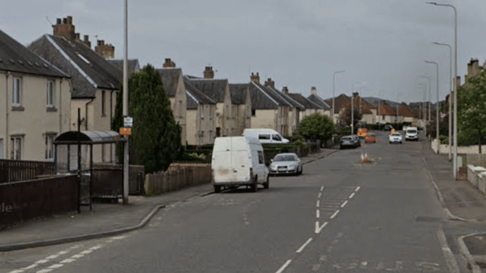 A road through a town. Houses are on either side and there is a bus stop on one side. Cars are parked on one side