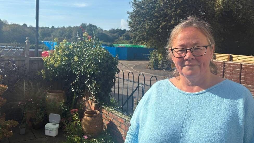A woman stood in her garden . She is wearing a blue top. In the background there is a blue barrier cordoning off a pumping station just up the road from her house.