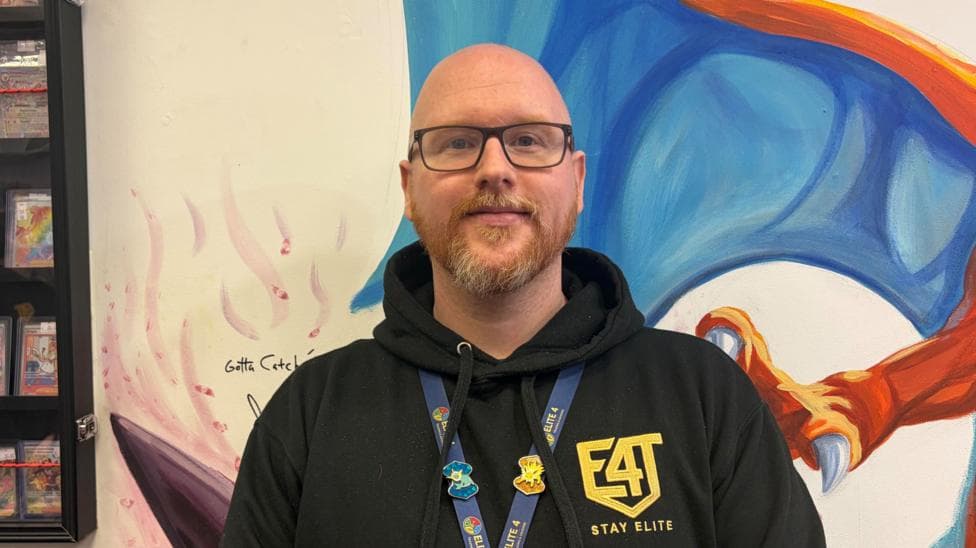 A bald man with a ginger beard. He is wearing glasses and has a black hoodie on with a gold logo.