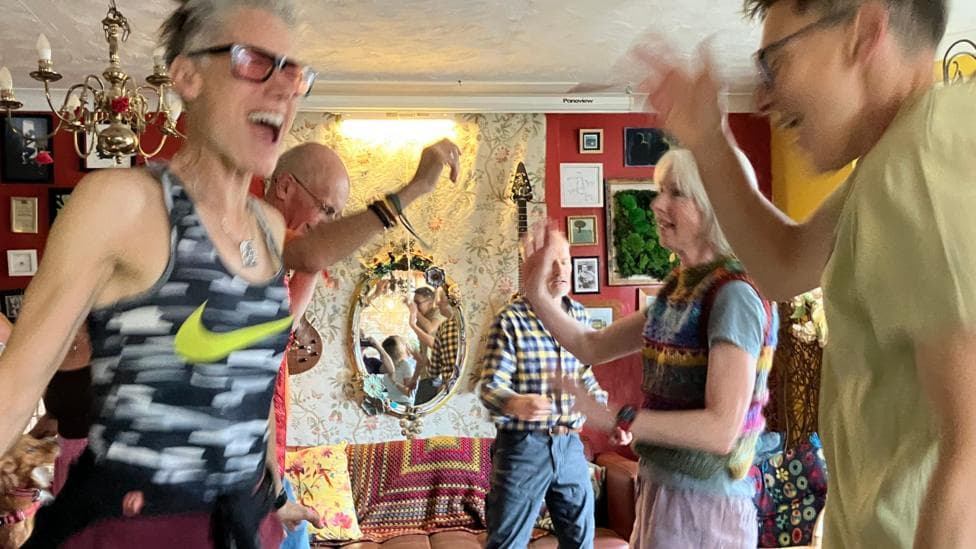A group of people are dancing in a café. There is a sofa in the background and a mirror on the wall. There are also pictures on the wall, which is painted red.